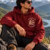 Mountain Is Calling Hoodie — Maroon — worn outdoors, seated The Mountain Is Calling hoodie in Maroon — worn outdoors, seated